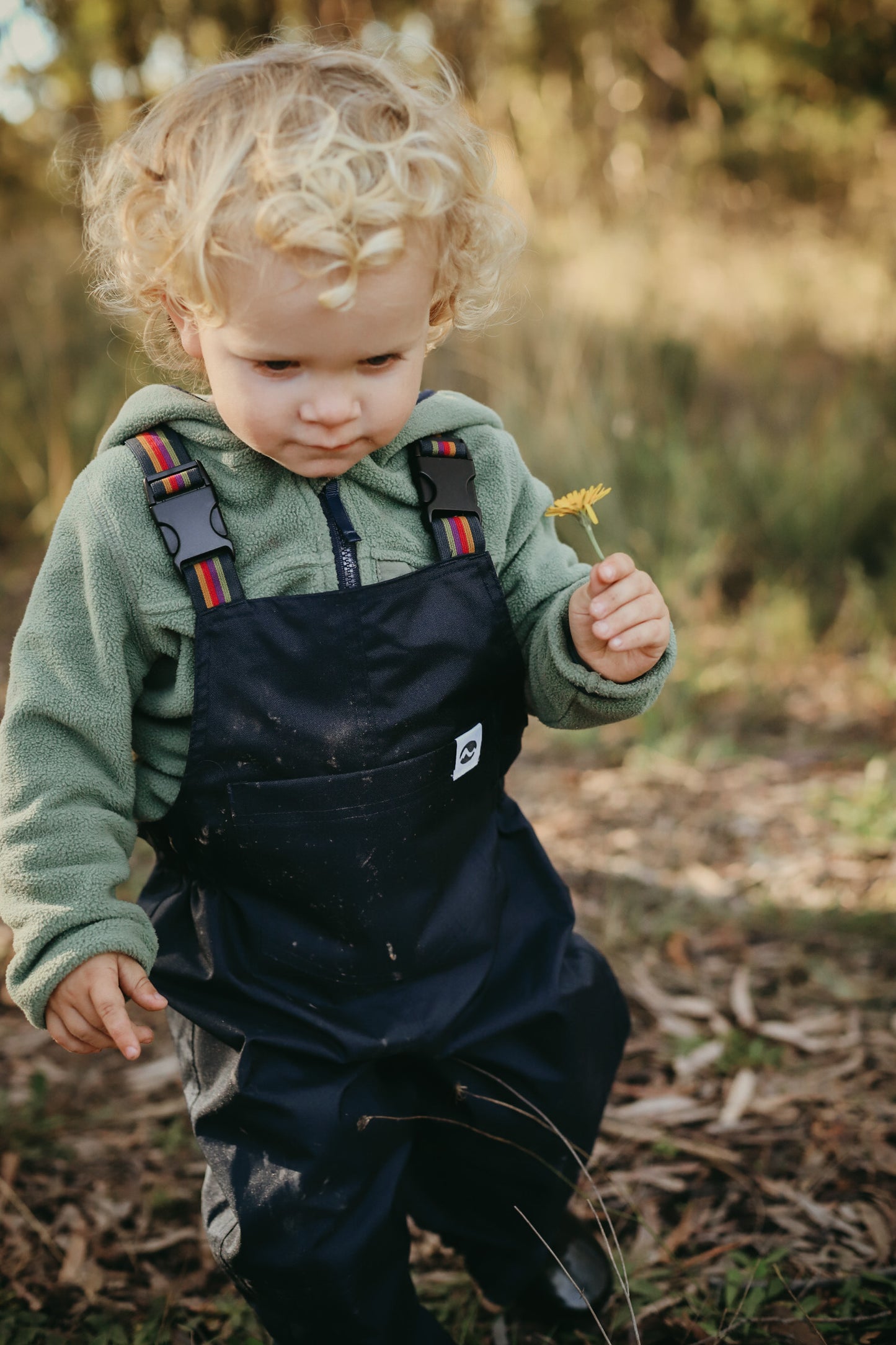 Child in navy waterproof overalls holding a flower outdoors