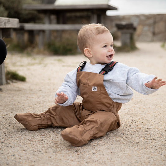 Child wearing brown overalls sitting on a gravel path with a blurred background