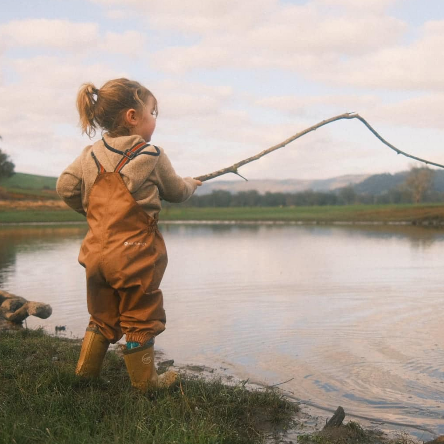 Child in brown waterproof overalls fishing by a lake with mountains in the background