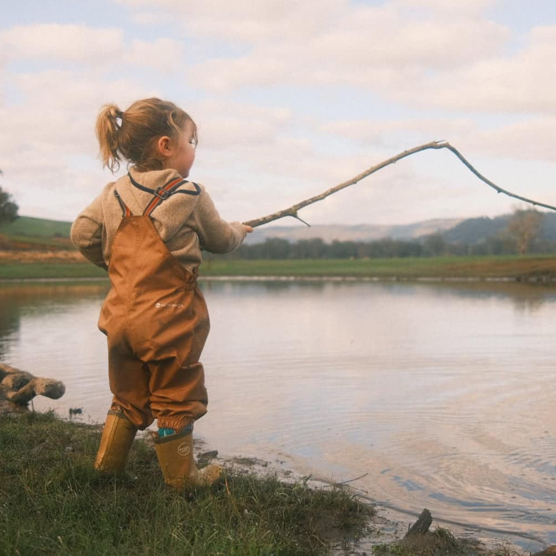 Child in brown waterproof overalls fishing by a lake with mountains in the background