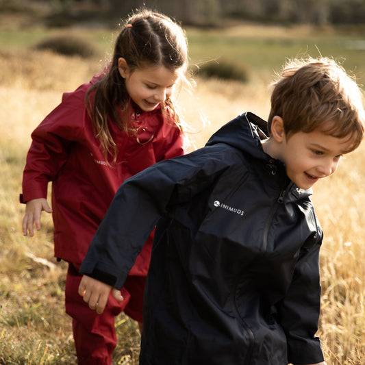 Two children in raincoats walking through a field with trees in the background